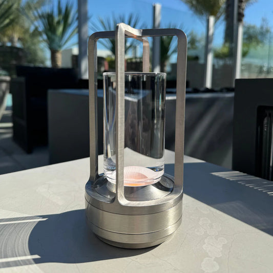 A modern, cylindrical LED lamp with a brushed metal frame and clear glass center, sitting on an outdoor table in bright sunlight. The lamp has a minimal, industrial design with vertical supports and a soft pink light at the base, surrounded by palm trees and patio furniture in the background.